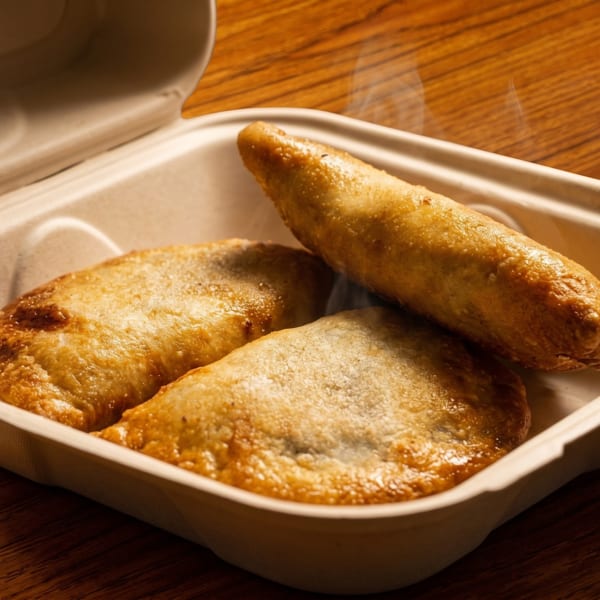 Three golden-brown, steaming empanadas in an open, eco-friendly takeout container on a wooden table.