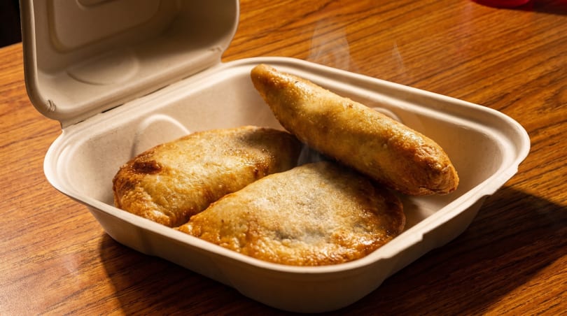 Three golden-brown, steaming empanadas in an open, eco-friendly takeout container on a wooden table.