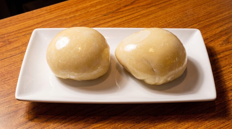 Two light-colored, smooth, round dumplings sit on a white rectangular plate on a wooden table.