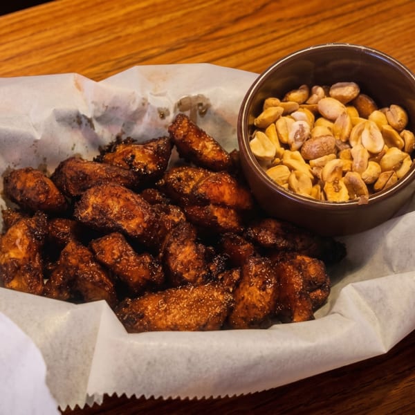 A basket of crispy chicken wings and a small bowl of peanuts on a wooden table.