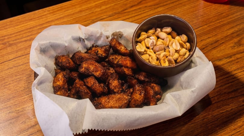 A basket of crispy chicken wings and a small bowl of peanuts on a wooden table.