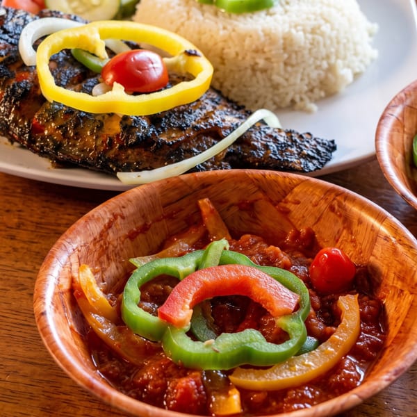 Grilled fish with rice, bell peppers, and onions, served with two bowls of stew.
