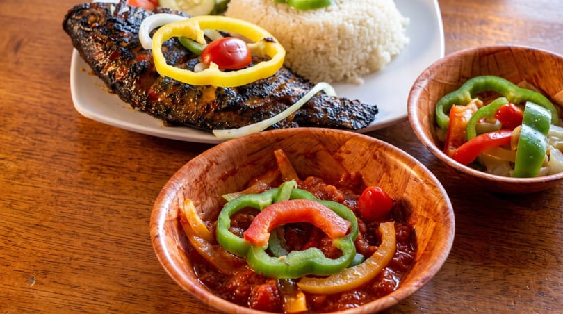 Grilled fish with rice, bell peppers, and onions, served with two bowls of stew.